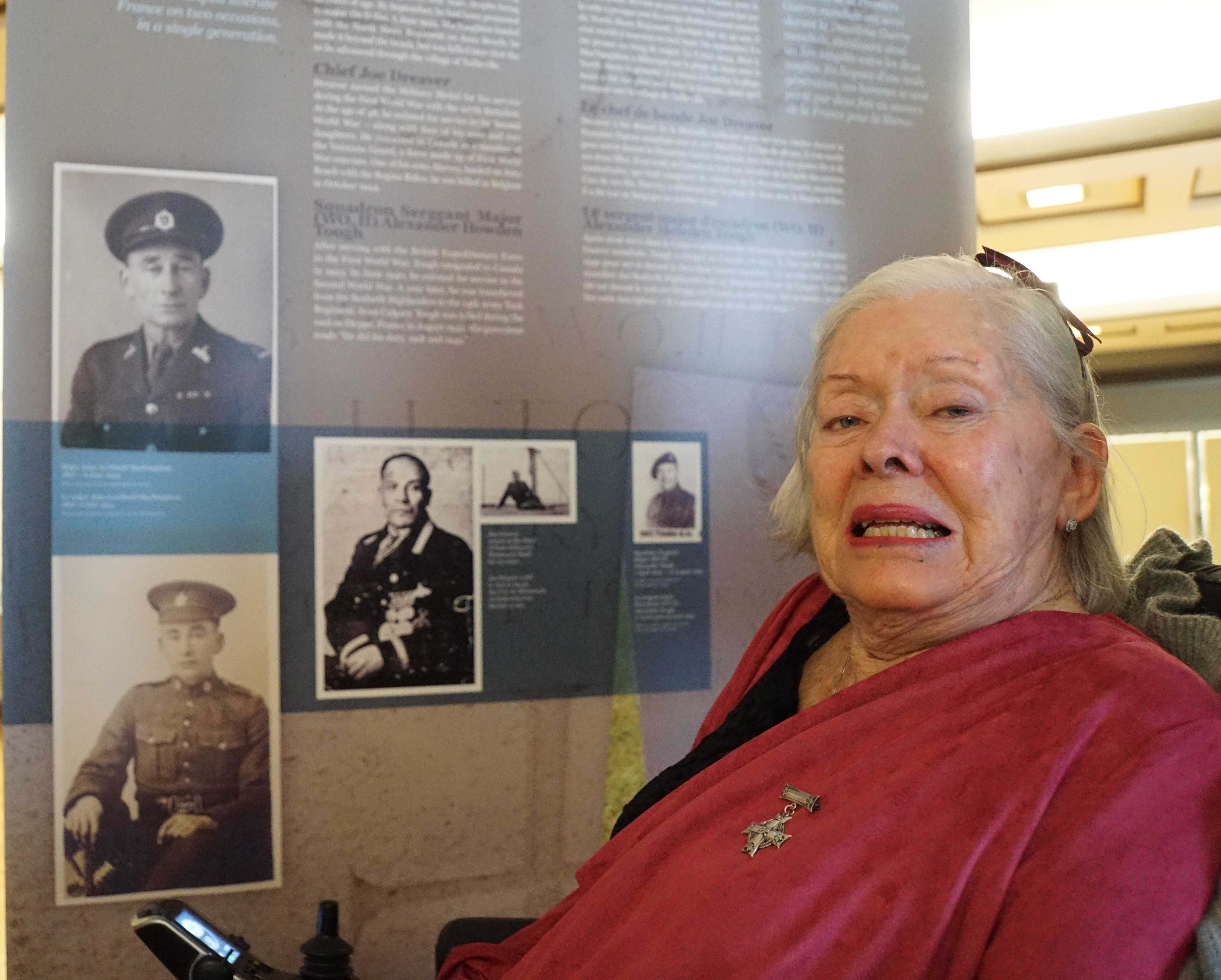 Colour photograph. Margie smiles in front of photos of her father, with her mother's Silver Cross pinned to her pink shawl